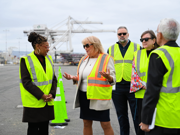 Image (Port of Oakland community stakeholders and officials in safety vests meeting on-site with port cranes in background discussing environmental initiatives)